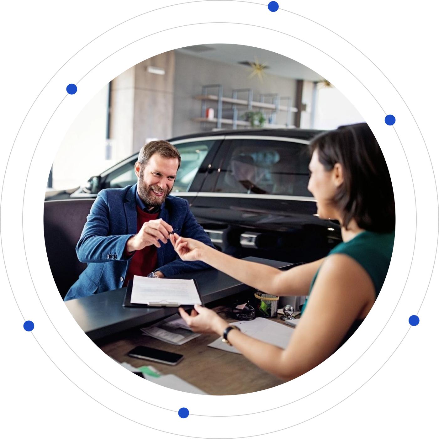 A man in a blue suit receives car keys from a woman at a dealership counter, with a black car in the background.