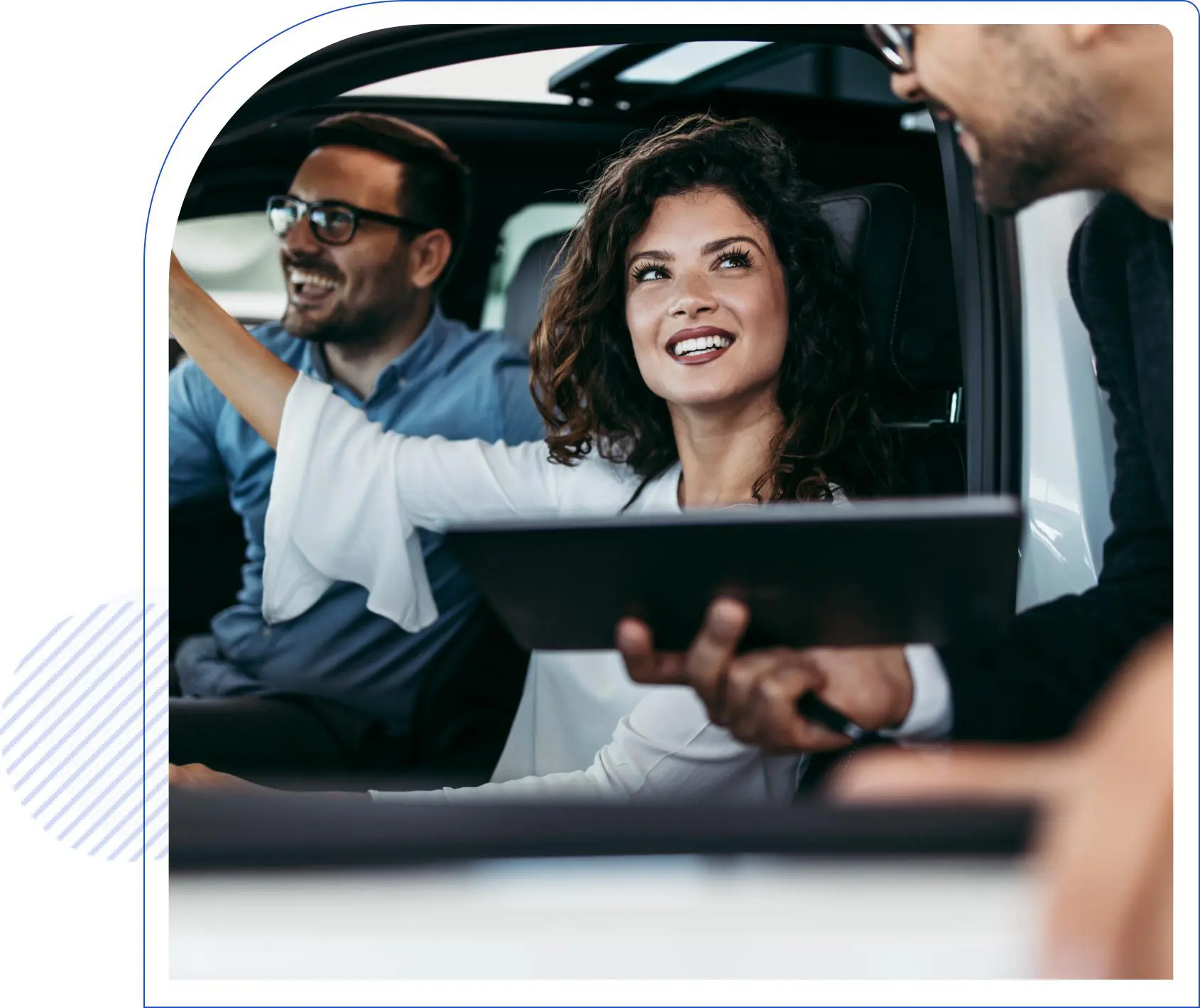 Two people inside a car are smiling and looking at a person holding a tablet outside the car window.