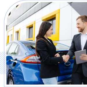 Man and woman shaking hands in front of a blue electric car, parked near a modern building. Woman holds car keys; man holds a tablet. Both are smiling.