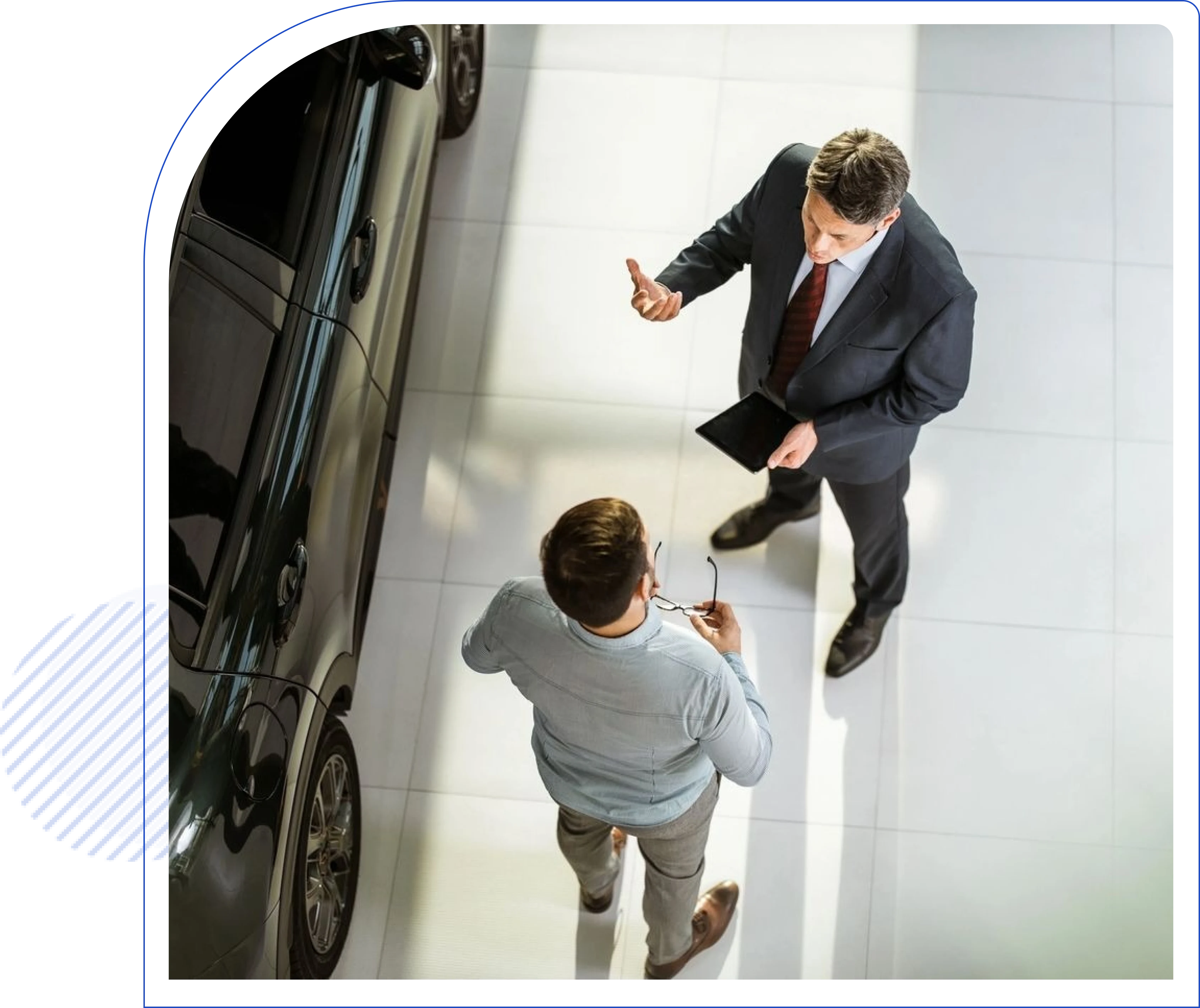 Two men stand indoors, talking next to a dark-colored car. One man holds a tablet while gesturing.