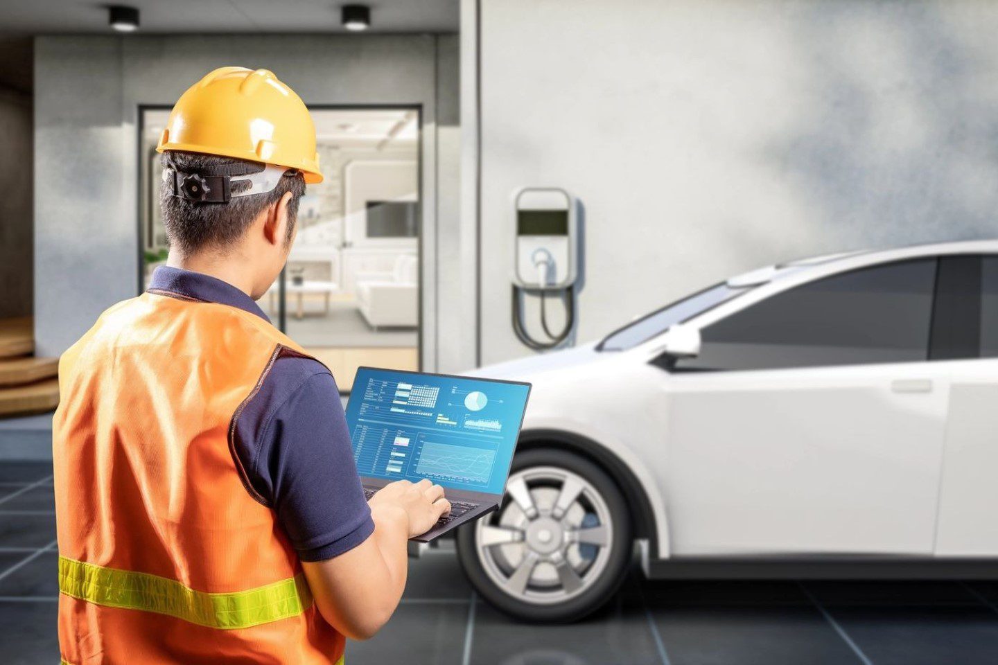 A person in a hard hat and safety vest uses a laptop near a white electric car parked by a charging station, with a building entrance in the background.