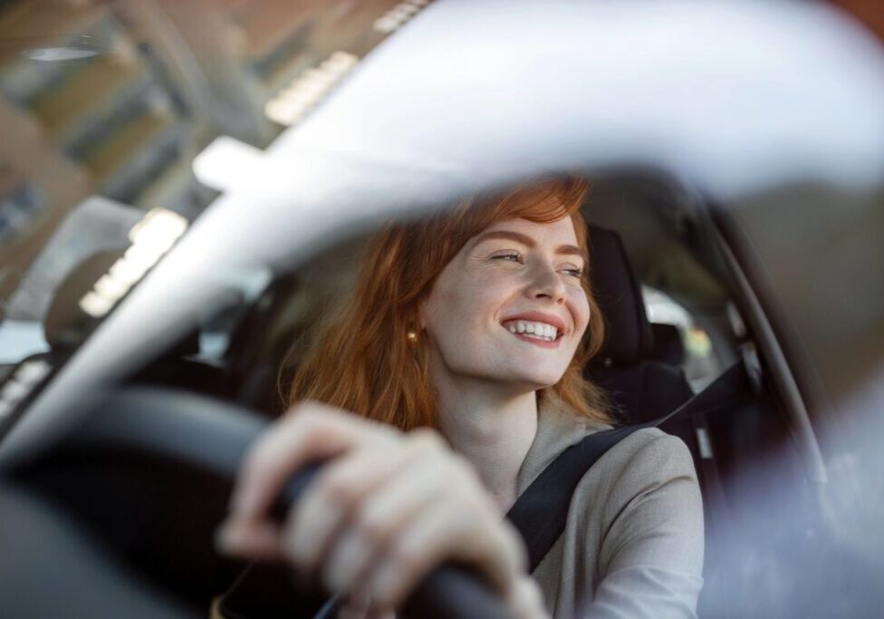 Woman smiling while driving a car, seen from the passenger side.