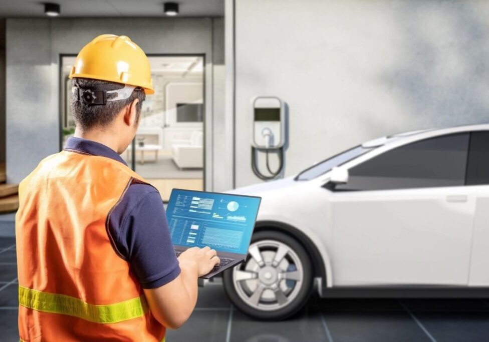 A person in a hard hat and safety vest uses a laptop near a white electric car parked by a charging station, with a building entrance in the background.