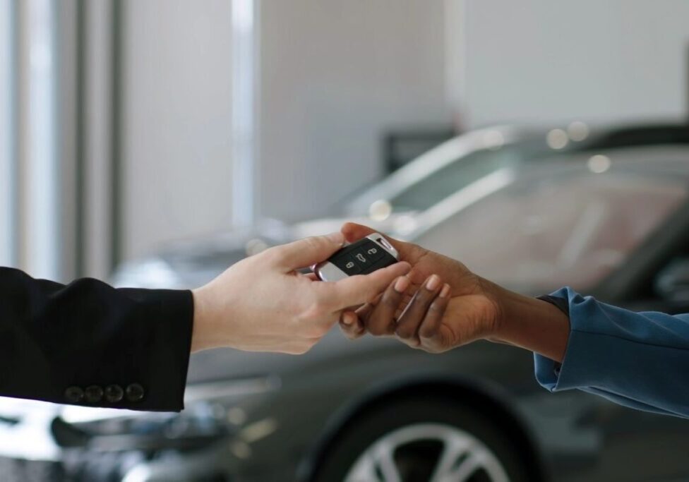 Two people exchanging a car key fob, with a blurred car in the background.